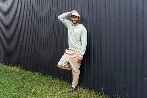 Longitud completa de hombre feliz en gorra de béisbol posando cerca de valla metálica - foto de stock