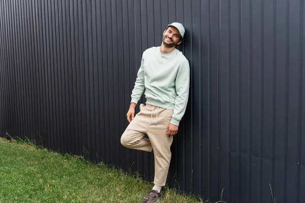 Full length of joyful man in baseball cap posing near metallic fence — Stock Photo
