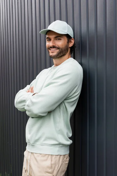 Gioioso uomo in berretto da baseball in posa con le braccia incrociate vicino recinto metallico — Foto stock