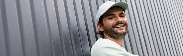 Low angle view of joyful man in baseball cap looking at camera near metallic fence, banner — Stock Photo