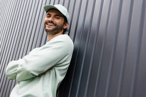 Low angle view of smiling man in baseball cap looking at camera near metallic fence — Stock Photo