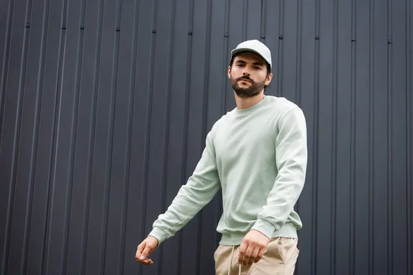 Hombre barbudo en gorra de béisbol y sudadera mirando a la cámara cerca de valla metálica - foto de stock