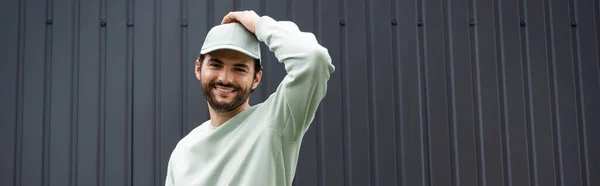 Smiling man in sweatshirt adjusting cap near metallic fence, banner — Stock Photo