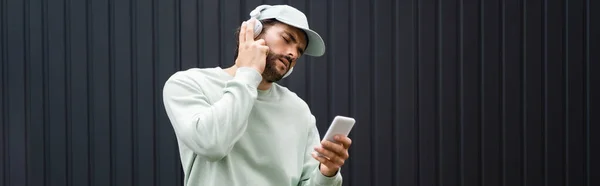 Bearded man in baseball cap listening music in wireless headphones near metallic fence, banner — Stock Photo