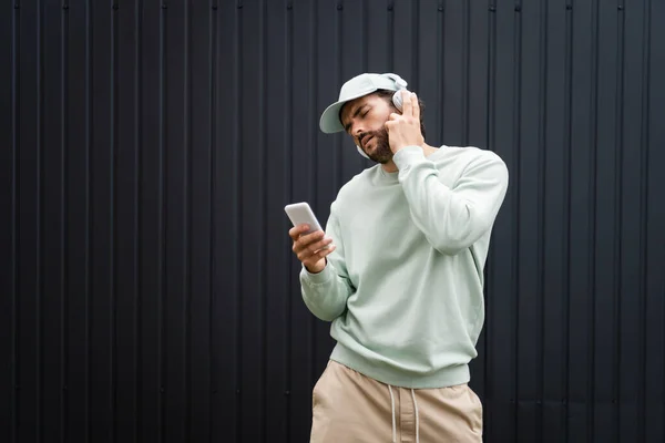 Bearded man in baseball cap listening music in wireless headphones near metallic fence — Stock Photo