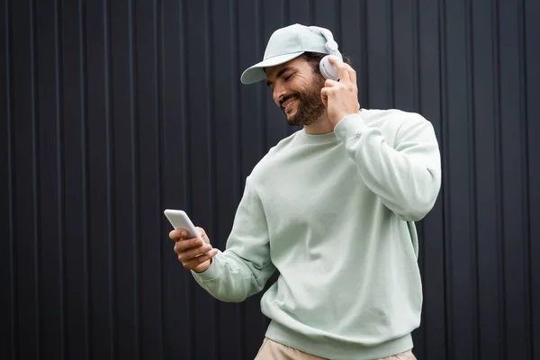 Happy man in baseball cap listening music in wireless headphones and holding smartphone near metallic fence — Stock Photo