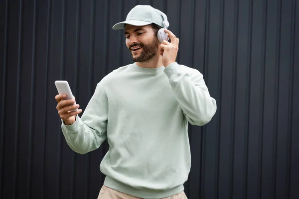 Fröhlicher Mann mit Baseballmütze, der mit drahtlosen Kopfhörern Musik hört und sein Smartphone in der Nähe eines Metallzauns hält — Stockfoto