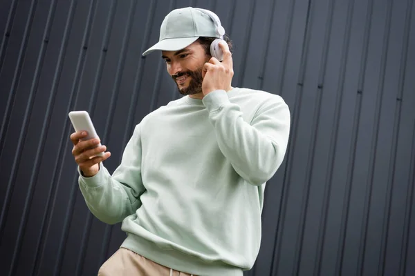 Homem positivo no cap ouvir música em fones de ouvido sem fio e segurando smartphone perto de cerca metálica — Fotografia de Stock