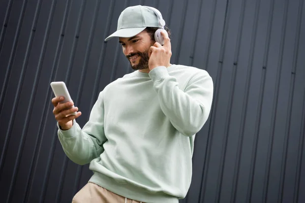 Positive man in cap listening music in wireless headphones and holding smartphone near metallic fence — Stock Photo