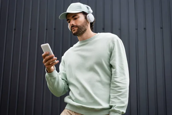 Bearded man in cap listening music in wireless headphones and holding smartphone near metallic fence — Stock Photo