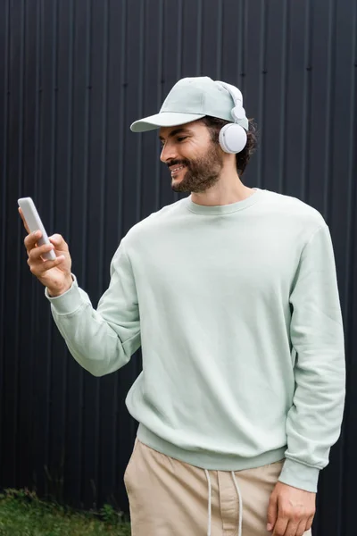 Positive man in cap listening music in wireless headphones and holding cellphone near metallic fence — Stock Photo