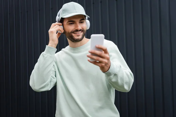 Positive man in cap listening music in wireless headphones and using cellphone near metallic fence — Stock Photo