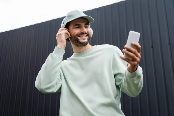 Visão de baixo ângulo do homem positivo no boné ouvir música em fones de ouvido sem fio e usando celular perto de cerca metálica — Fotografia de Stock