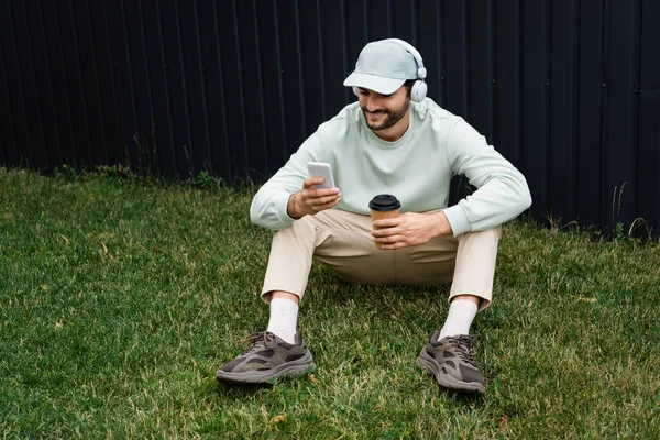 Positiver Mann mit Mütze hört Musik in drahtlosen Kopfhörern und benutzt Handy, während er auf grünem Rasen in der Nähe von Zaun sitzt — Stockfoto