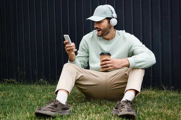 Erstaunter Mann mit Mütze hört Musik in drahtlosen Kopfhörern und benutzt Handy, während er auf grünem Rasen in der Nähe von Zaun sitzt — Stockfoto