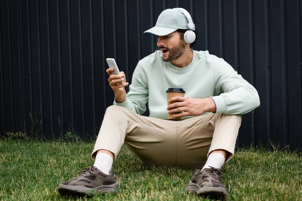 Amazed man in cap listening music in wireless headphones and using cellphone while sitting on green lawn near fence — Stock Photo