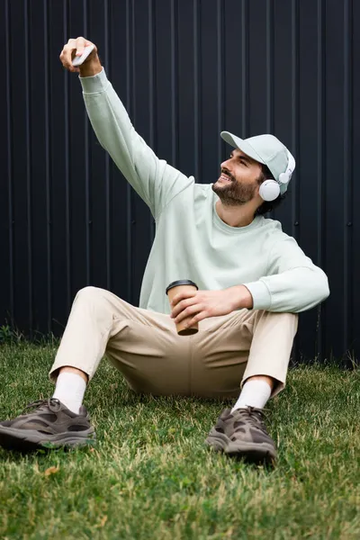 Fröhlicher Mann mit Schirmmütze und kabellosen Kopfhörern, der auf grünem Rasen mit Pappbecher ein Selfie macht — Stockfoto