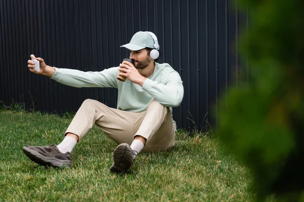 Веселий чоловік в бездротових навушниках приймає селфі, сидячи на зеленому газоні з паперовою чашкою — стокове фото