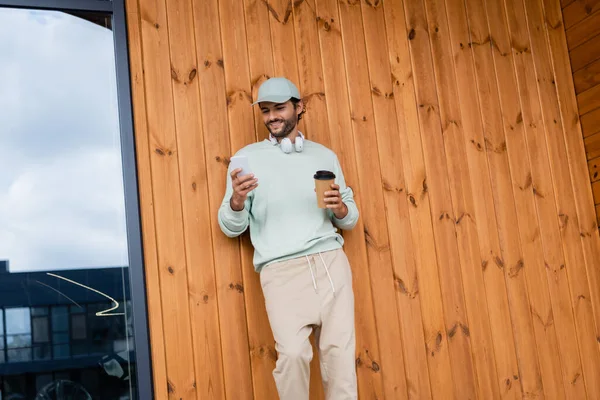 Позитивный человек в кепке и беспроводных наушников с помощью мобильного телефона, держа бумажную чашку возле здания — стоковое фото