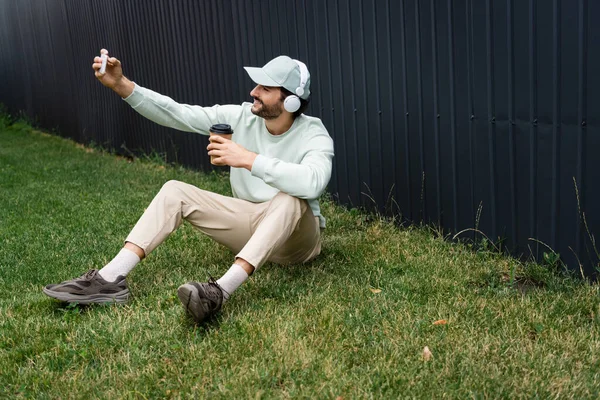 Hombre positivo en auriculares inalámbricos tomando selfie mientras está sentado en hierba verde con taza de papel - foto de stock