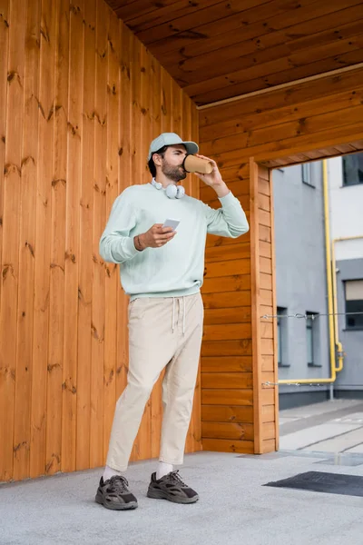 Homme barbu en chapeau boire du café à emporter tout en tenant smartphone près du bâtiment — Photo de stock