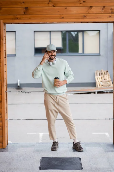 Volle Länge des glücklichen Mannes mit Mütze, der Coffee to go in der Hand hält, während er in der Nähe des Gebäudes mit dem Smartphone spricht — Stockfoto