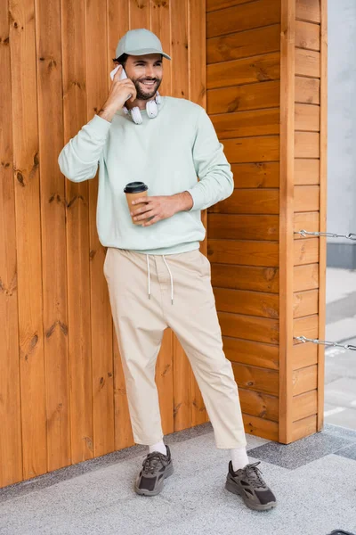 Piena lunghezza di uomo allegro in berretto da baseball che tiene il caffè per andare mentre si parla su smartphone vicino edificio — Foto stock