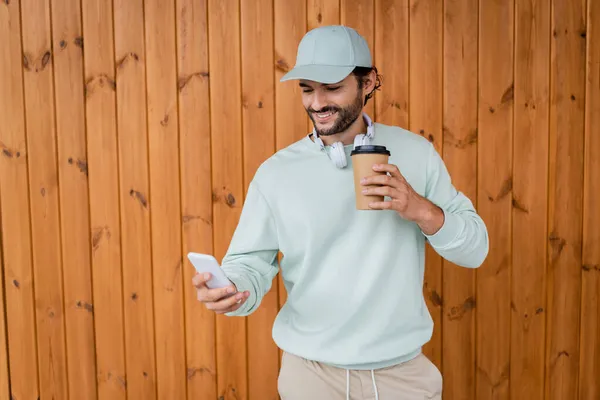 Щасливий чоловік у кепці тримає каву, щоб піти під час використання смартфона — стокове фото