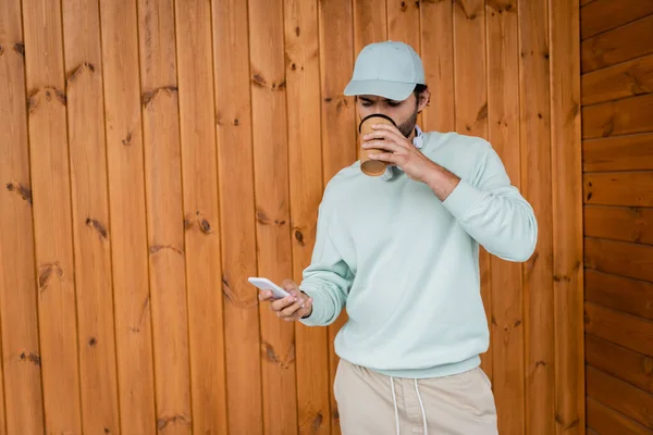 Bearded man in cap drinking coffee to go while using smartphone — Stock Photo