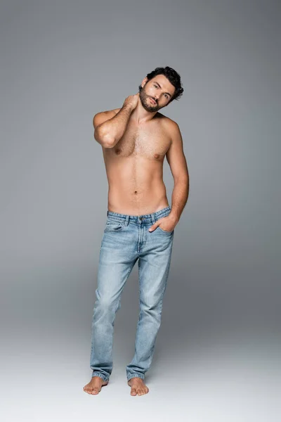 Full length of good-looking and muscular man in blue jeans posing with hand in pocket on grey — Stock Photo