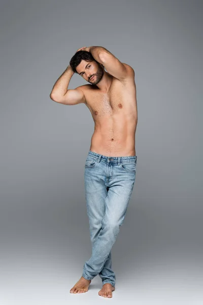 Comprimento total de homem bonito e musculoso em jeans olhando para a câmera enquanto posando em cinza — Fotografia de Stock