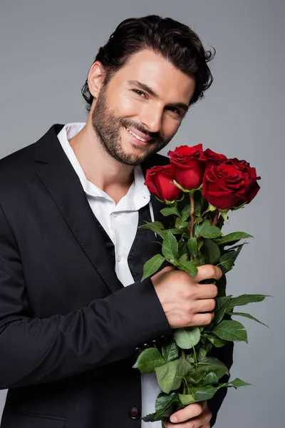 Hombre positivo en traje sosteniendo ramo de rosas rojas aisladas en gris - foto de stock