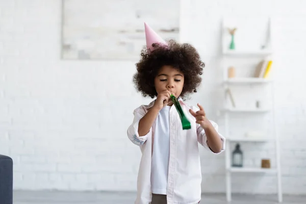 African american kid in party cap blowing in party horn in living room — Stock Photo