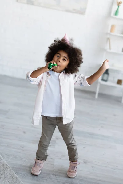 Full length view of happy african american birthday girl blowing in party horn at home — Stock Photo