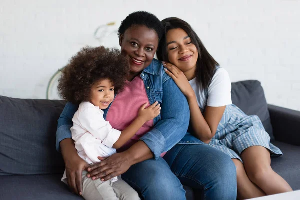 Femme américaine africaine mature souriant à la caméra tout en embrassant sur le canapé avec la jeune fille et l'enfant en bas âge — Photo de stock