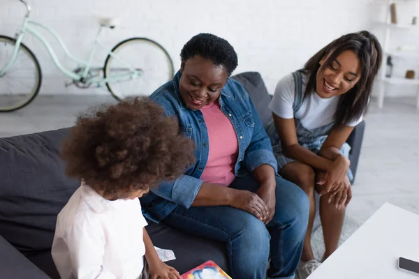 Alegres mujeres afroamericanas mirando a la chica rizada sentada en el sofá con libro de imágenes - foto de stock