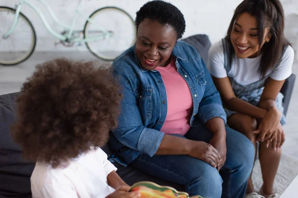 Curly african american child with picture book sitting on sofa with happy mom and granny — Stock Photo