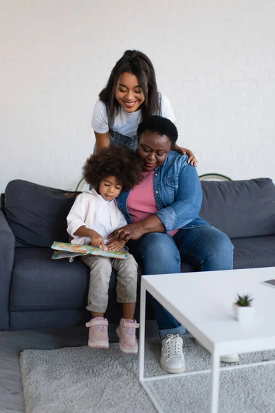 African american girl with granny pointing at picture book while sitting on sofa near smiling mom — Stock Photo