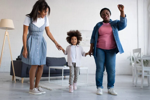 Cheerful african american girl holding hands with mother and granny while dancing together in living room — Stock Photo
