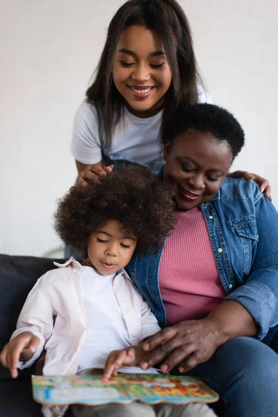 African american toddler girl looking at picture book near granny and happy mother — Stock Photo