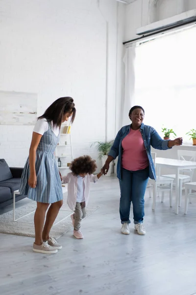 Alegres mujeres afroamericanas cogidas de la mano y bailando con una niña en casa - foto de stock