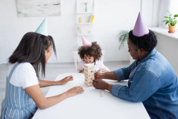 Felice ragazza afroamericana in berretto partito giocare blocchi di legno gioco con la mamma e la nonna in berretti partito — Foto stock