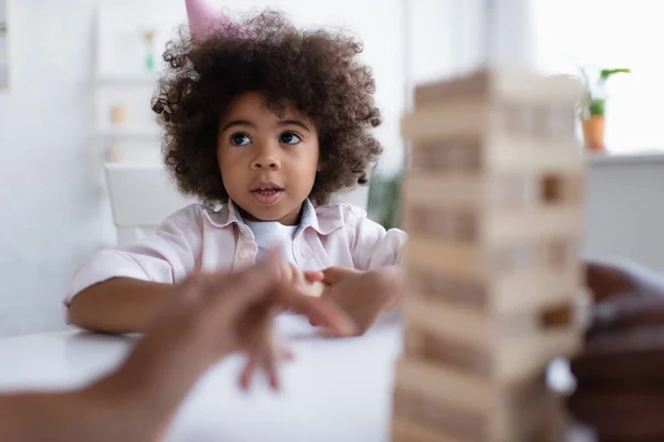 Lockige afrikanisch-amerikanische Mädchen in Partymütze Blick auf verschwommene Familie spielen Holzklötze Spiel — Stockfoto