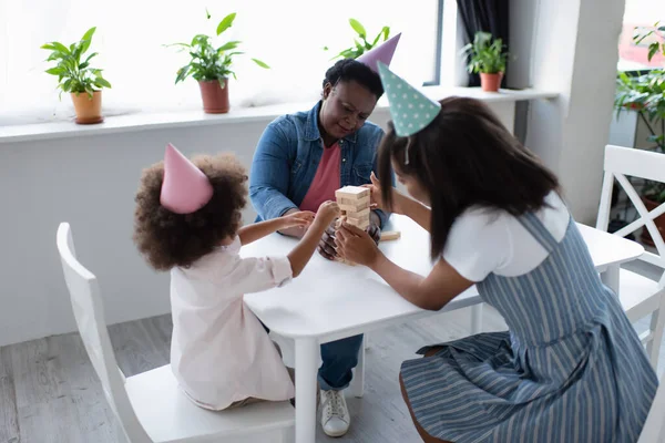 Afro-américain gosse avec mère et mamie en casquettes de fête jouer jeu de blocs de bois à la maison — Photo de stock