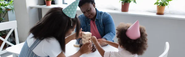African american women and toddler child in party caps playing wood blocks game at home, banner — Stock Photo