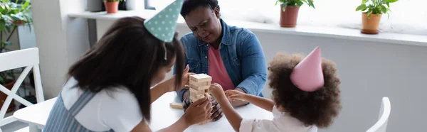 Mujeres afroamericanas y niños pequeños en gorras de fiesta jugando bloques de madera juego en casa, pancarta - foto de stock