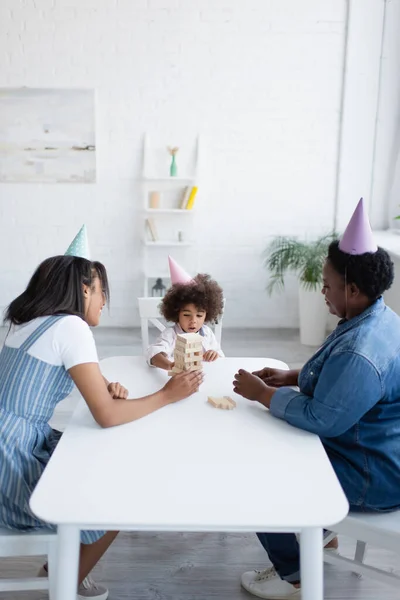 Mujeres afroamericanas y niña en gorras de fiesta jugando bloques de madera juego en casa - foto de stock