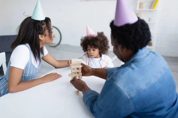 Afro-américaine fille avec mère souriante et mamie floue dans les casquettes de fête jouer jeu de blocs de bois — Photo de stock