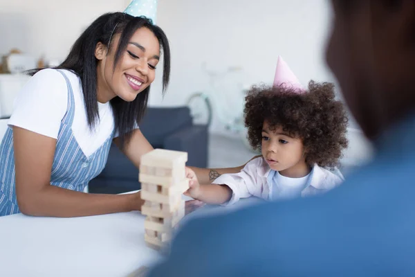 Souriant afro-américaine femme et fille en casquettes de fête jouer jeu de blocs de bois près de mamie floue — Photo de stock