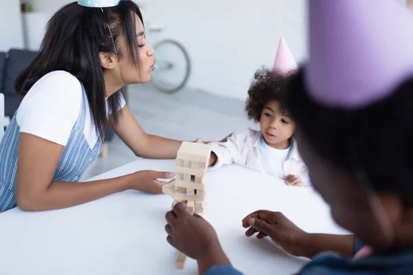 Blurred african american woman playing wood blocks game with daughter and granddaughter in party caps — Stock Photo